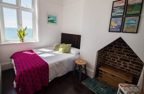 Ein Schlafzimmer mit Bett, einem Fenster mit Aussicht, einem kleinen Hocker, einem Kamin aus Ziegelsteinen und gerahmten Bildern an der Wand.