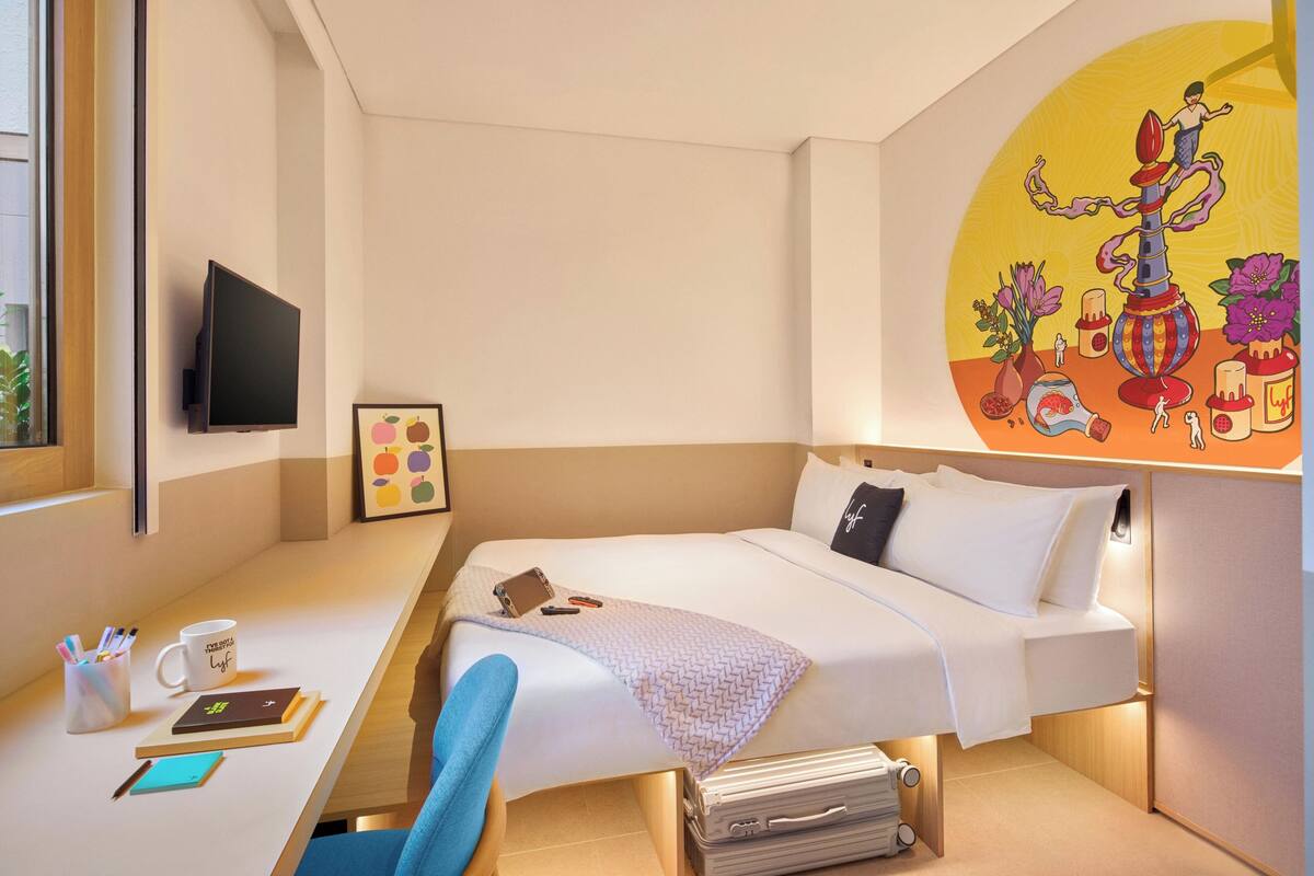 Een hotelkamer met een bed, nachtkastje, stoel, televisie, kunstwerk en een koffer.