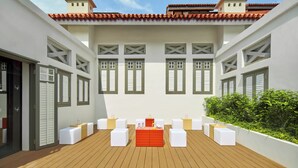 Um pátio com deck de madeira, assentos vermelhos e brancos, e um edifício branco com persianas verdes.