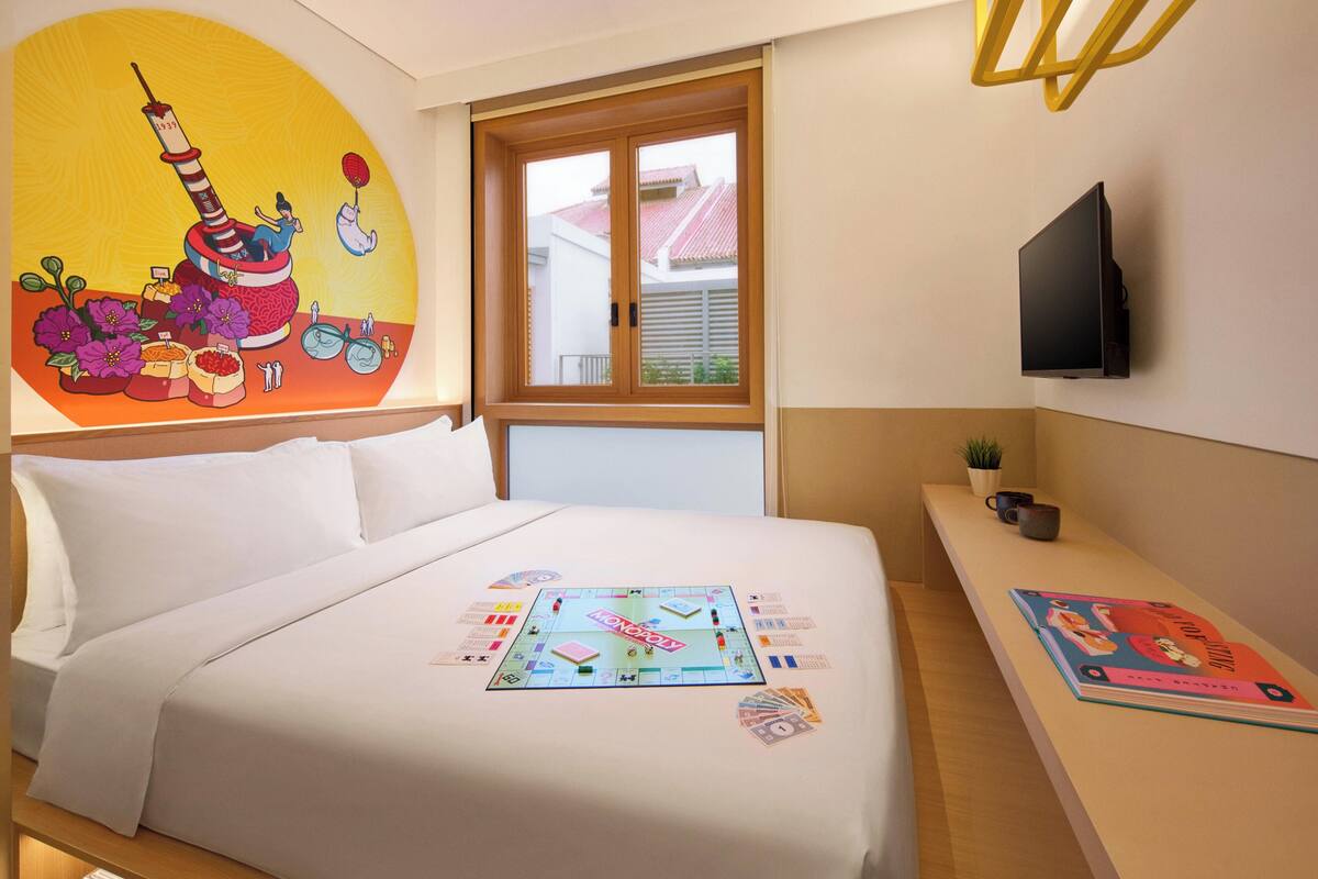 Een hotelkamer met een bed, nachtkastje, televisie, raam en een schilderij aan de muur.