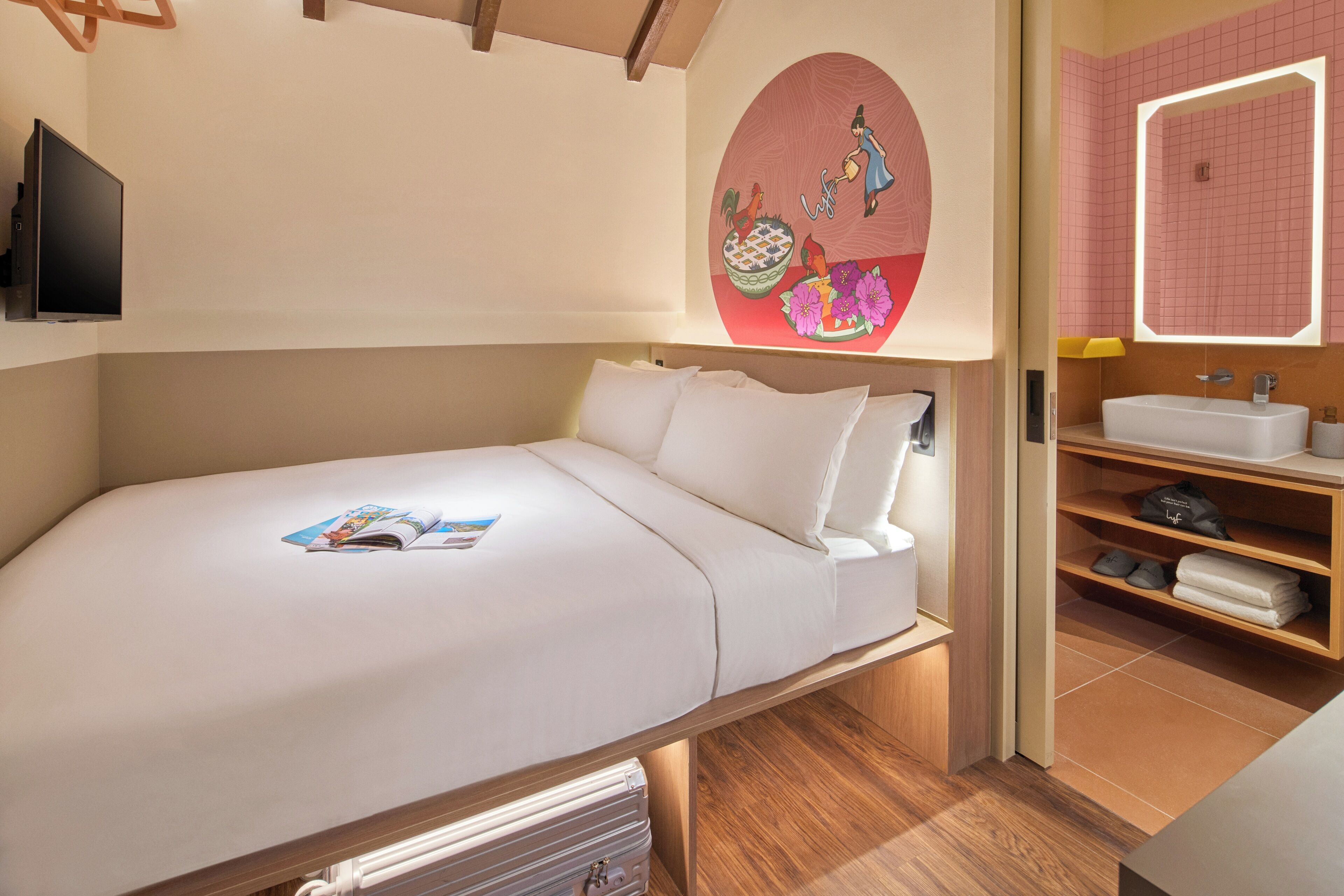 Une chambre à coucher comprenant un lit, un téléviseur, une œuvre d’art murale, une salle de bain avec un lavabo et un miroir, et un plancher en bois.