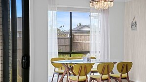 A dining area with a glass table, yellow chairs, a chandelier, and a view of a wooden fence outside.