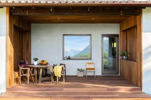 A wooden deck with a table and chairs, a window with a mountain view, and a door leading inside.