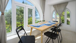 Un coin repas avec une table dressée pour quatre personnes, de grandes fenêtres avec des rideaux et une vue sur la verdure et un plan d’eau.