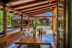 A covered outdoor seating area with wooden benches and tables, a green vase with flowers, and a view of a traditional building in the background. - Family Retreat Near Zagreb With Pool & Garden (Velika Gorica)