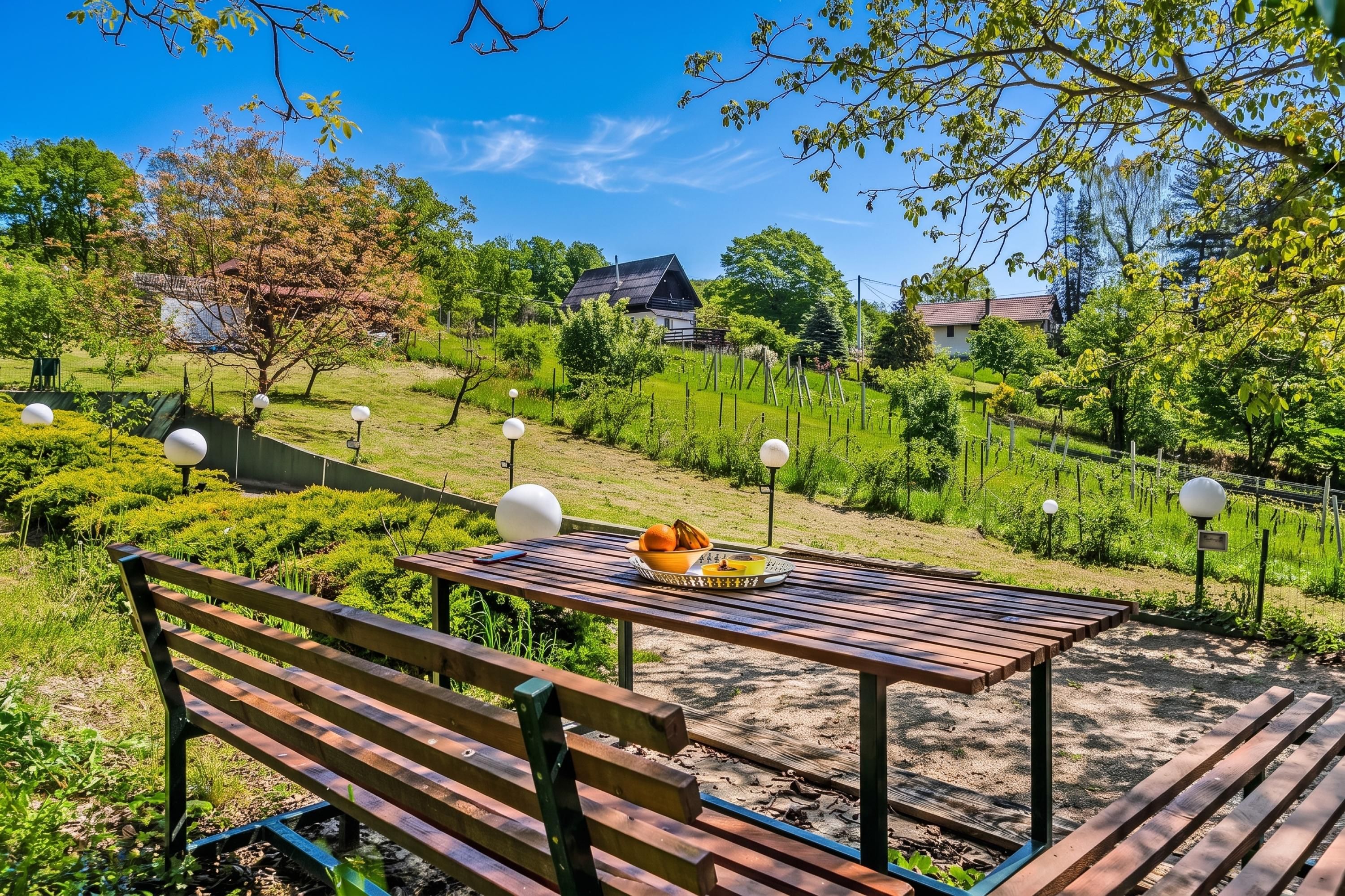 A wooden bench with a table in a garden setting, surrounded by greenery and a clear sky.