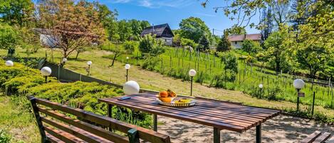 A wooden bench with a table in a garden setting, surrounded by greenery and a clear sky.