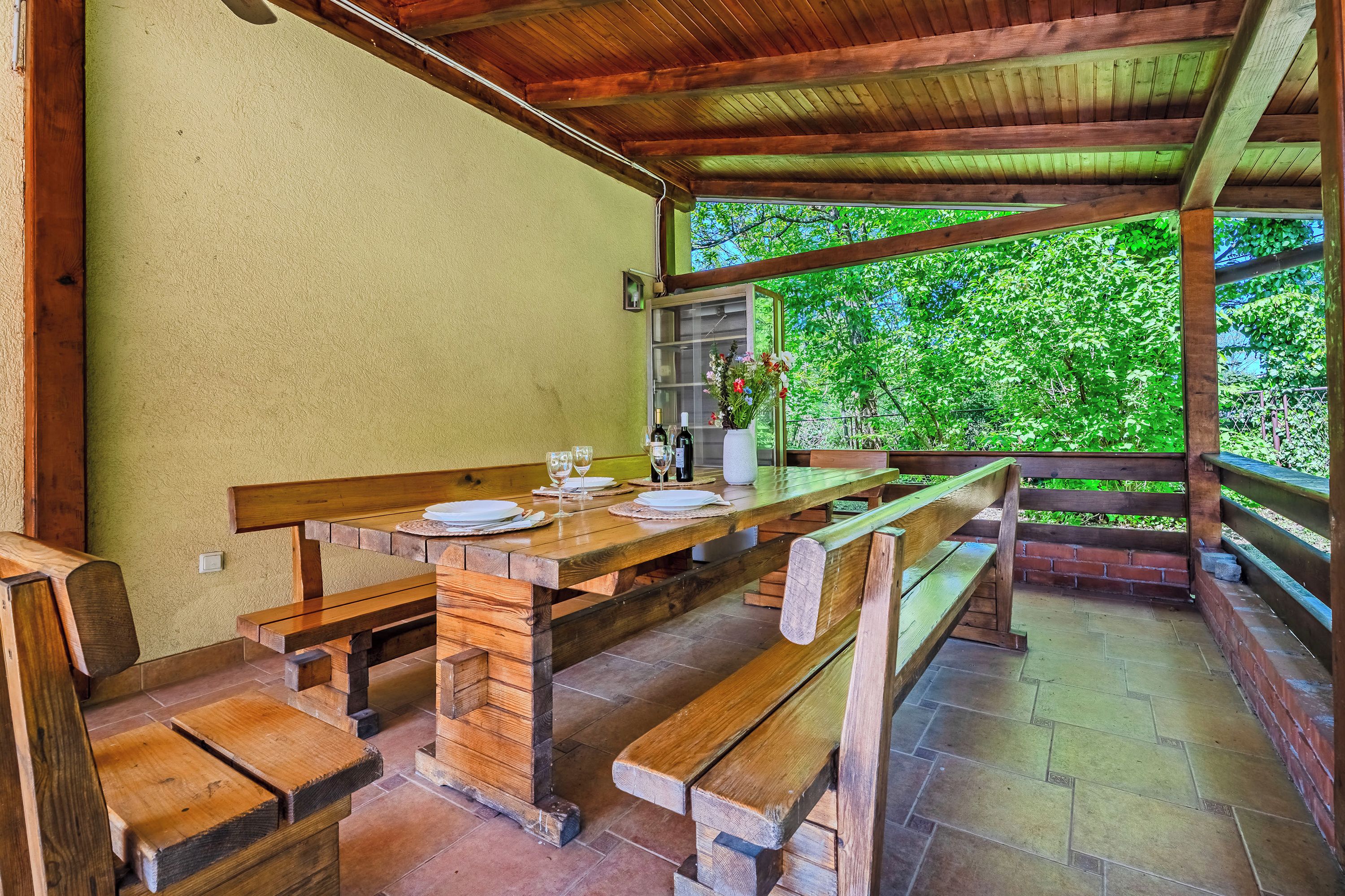 Una mesa de madera al aire libre con bancos, una planta en maceta y una ventana con vistas a la vegetación.