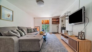 Moderni olohuone, jossa on harmaa kulmasohva, puinen konsolipöytä, litteänäyttöinen televisio ja suuri ikkuna, josta avautuu näkymä ulos.