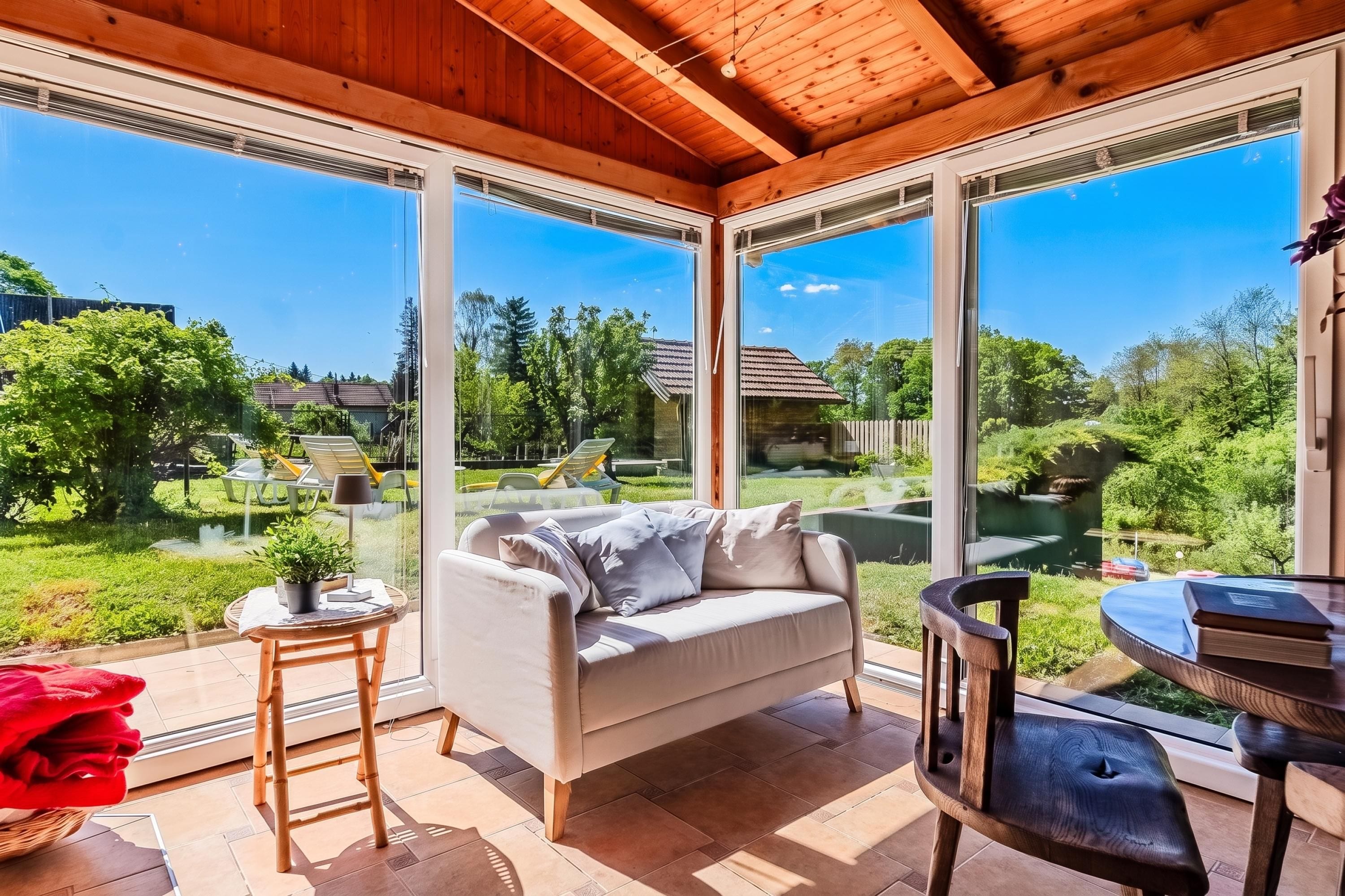 Ein sonnendurchflutetes Zimmer mit einer Couch, einem Tisch und einem Sessel, große Fenster mit Blick auf einen Garten und Häuser.