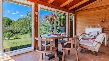 Interior de madera con una mesa redonda, sillas y vista a un jardín a través de grandes ventanales.