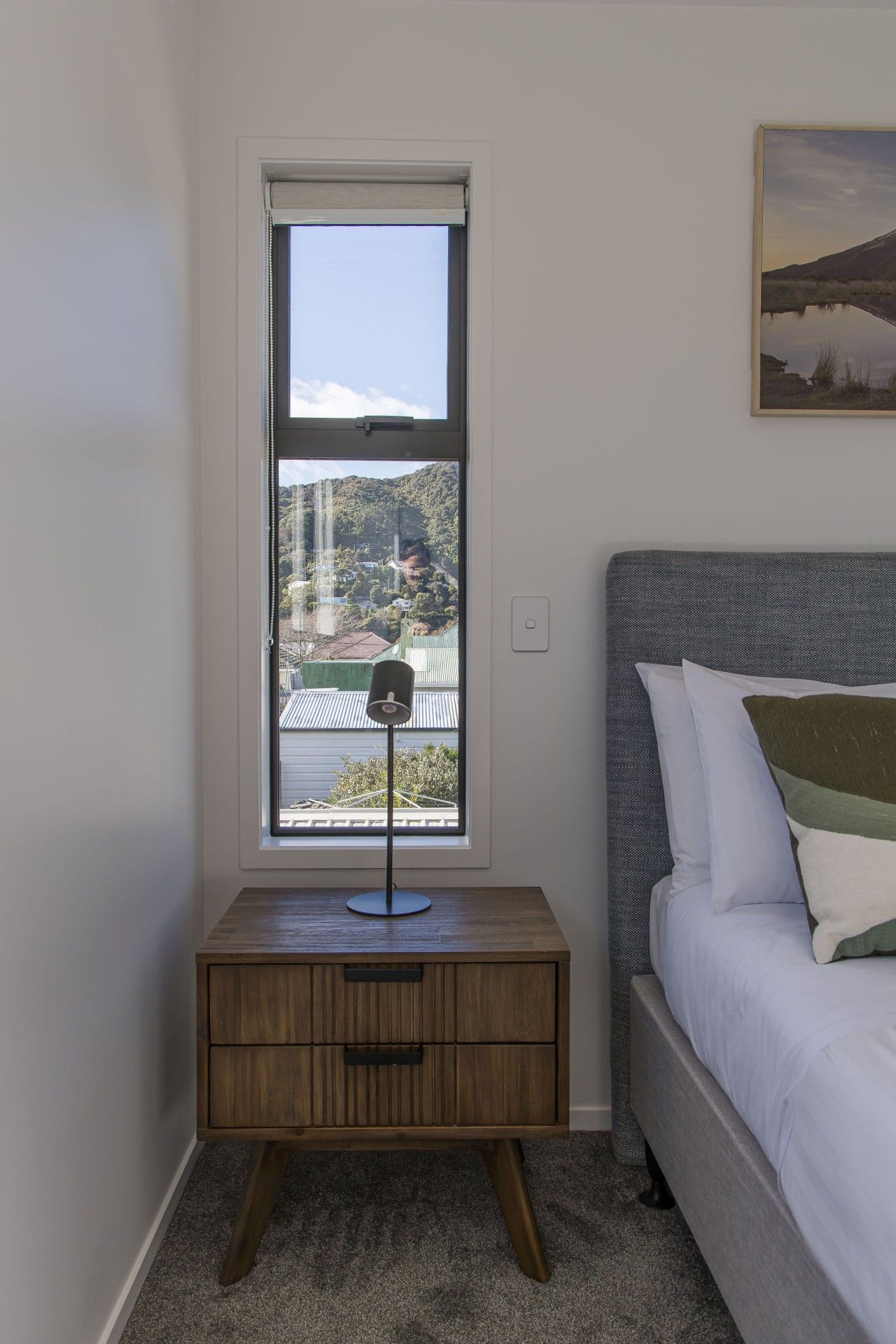 A bedroom with a bed, a nightstand with a lamp, a window with a view of a landscape, and a painting on the wall.