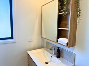 Une salle de bain avec un lavabo blanc, un miroir, une étagère en bois et une plante.