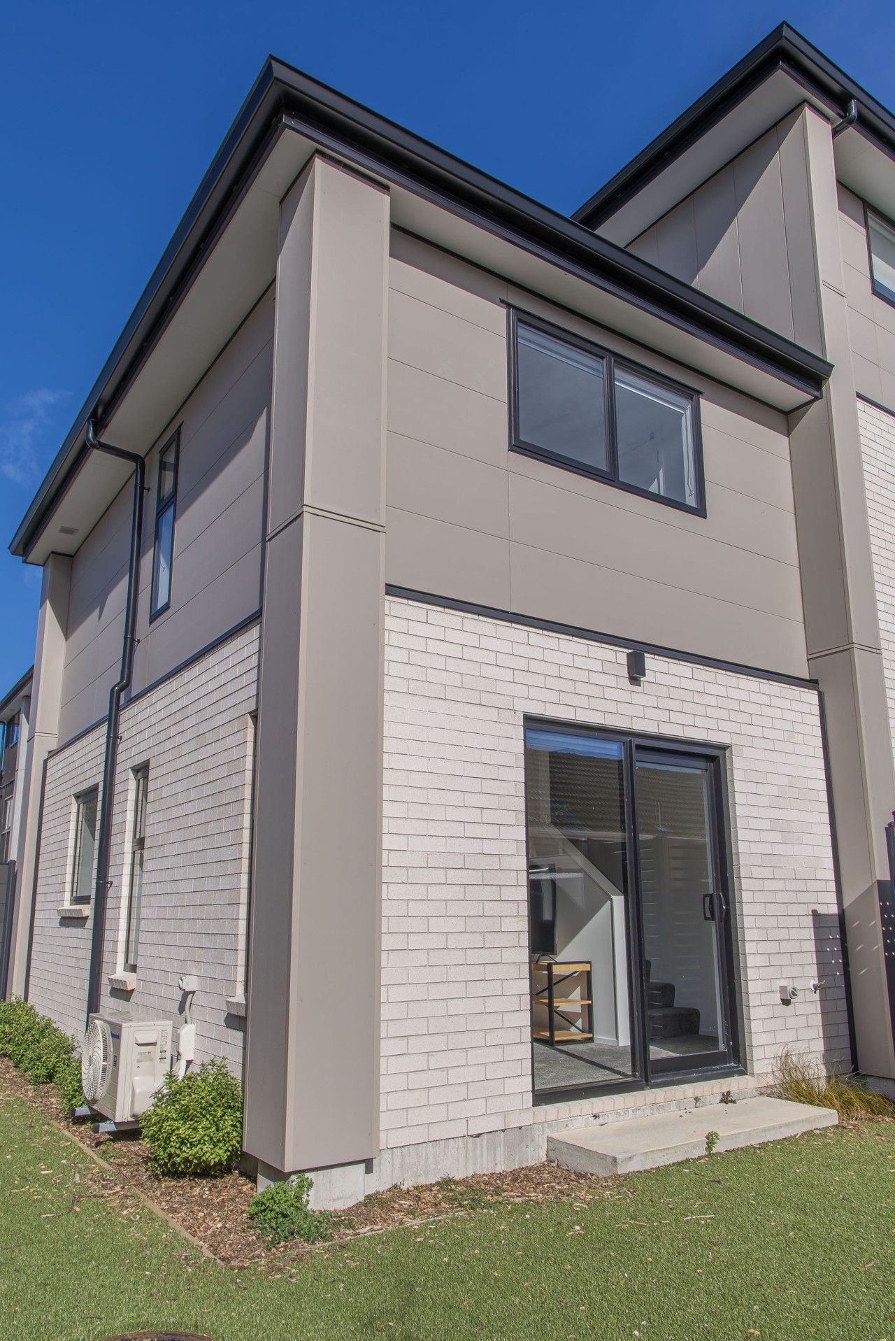 A modern two-story residential building with a brick facade, large sliding glass doors, and a visible air conditioning unit.