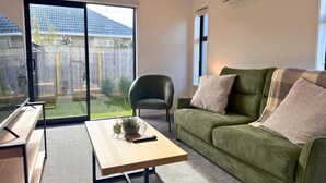 Un salon avec un canapé vert, une table basse en bois, un fauteuil vert et une porte-fenêtre coulissante donnant sur un jardin.