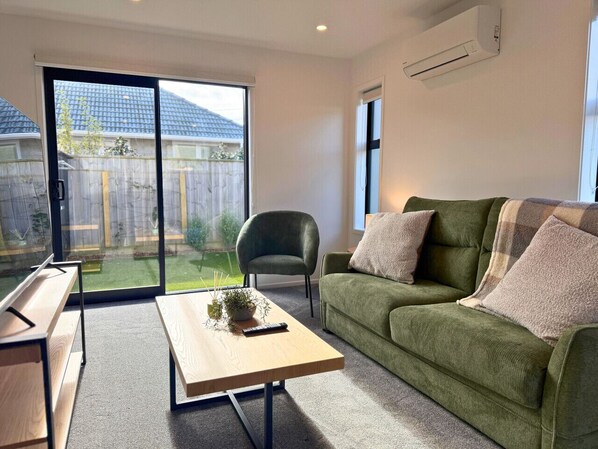 Un salon avec un canapé vert, une table basse en bois, un fauteuil vert et une porte-fenêtre coulissante donnant sur un jardin.
