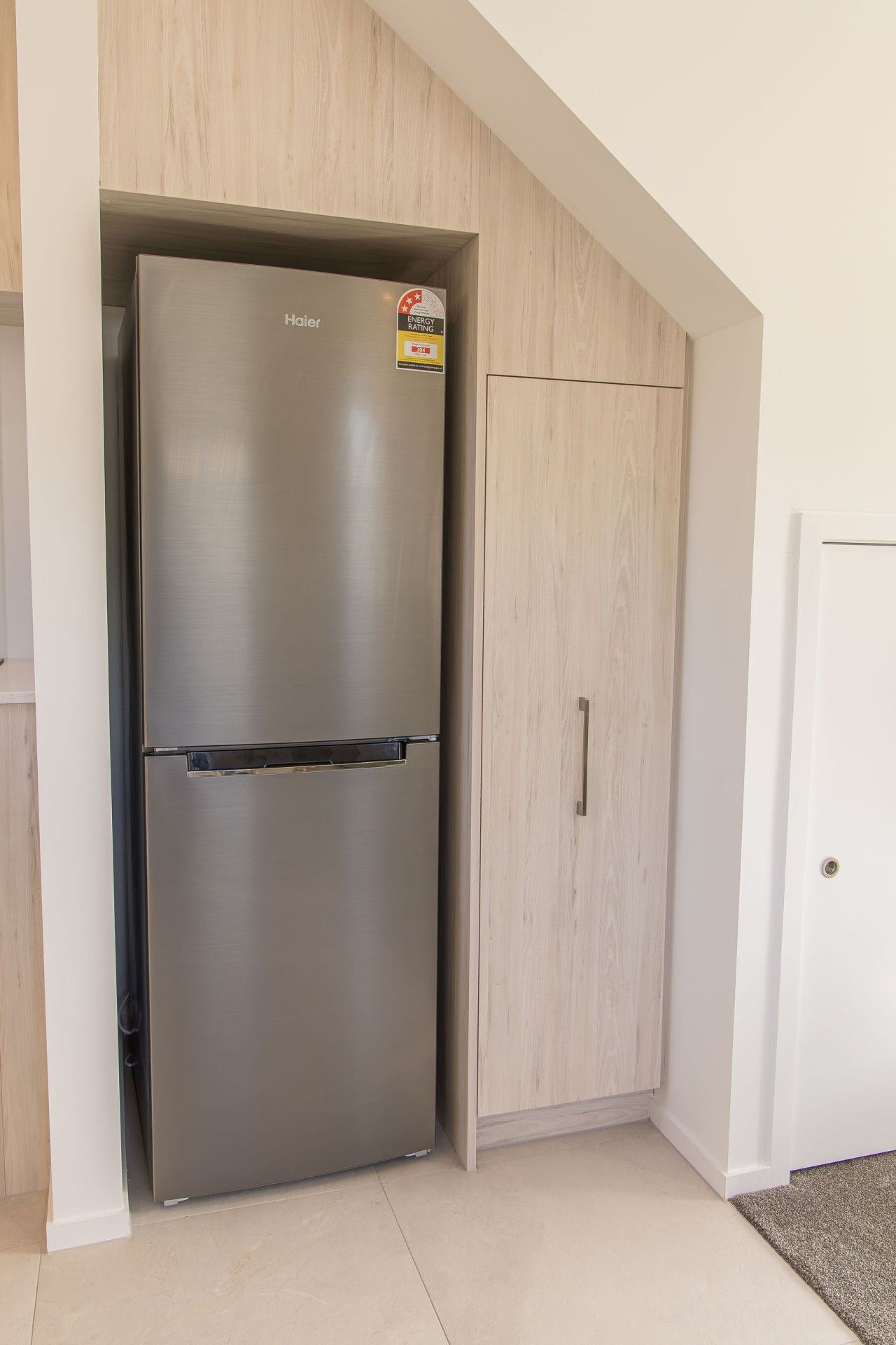 A stainless steel refrigerator with a sticker indicating an energy rating, positioned next to a wooden wardrobe in a room with white walls and a tiled floor.