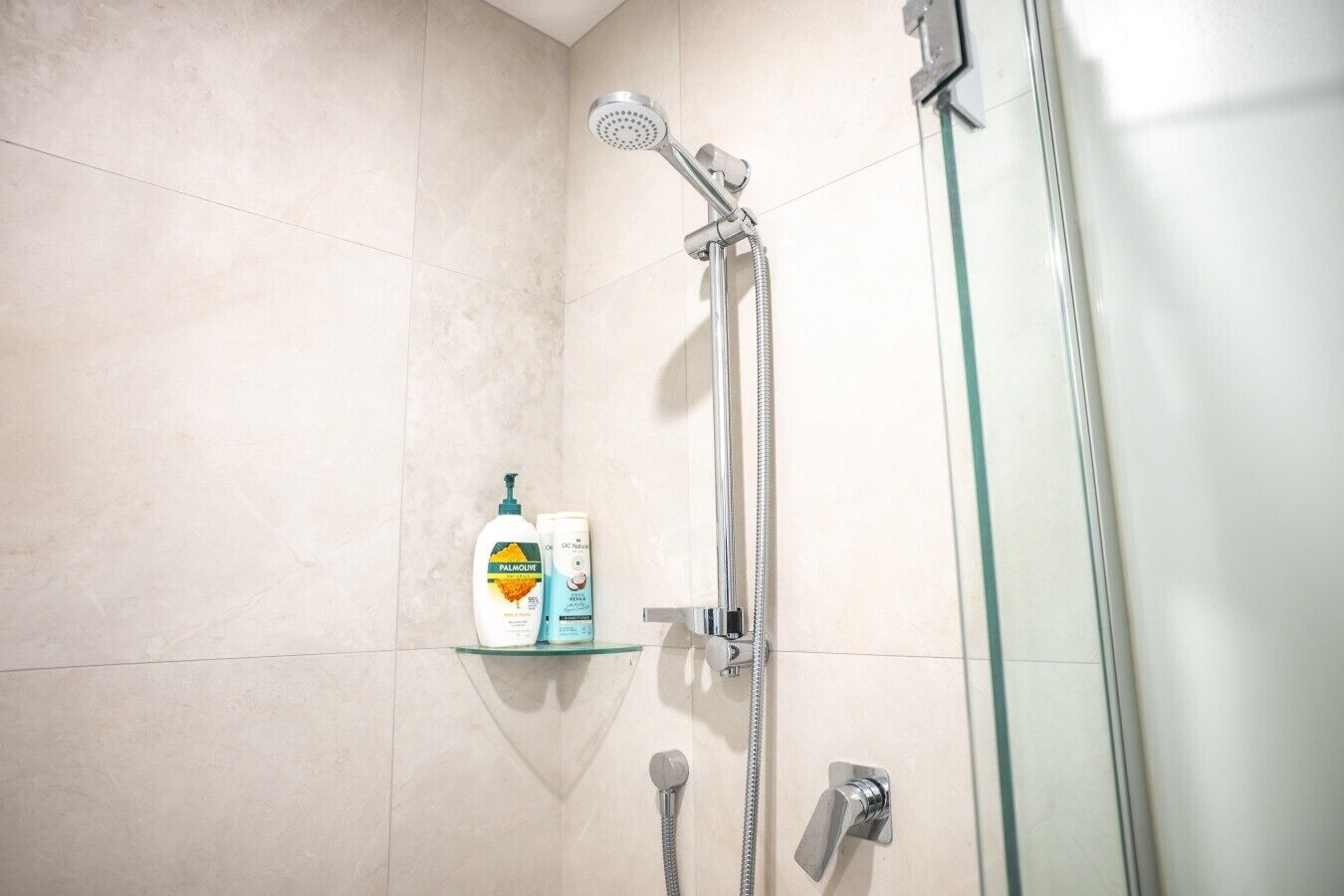 Een douche met een handdouche en een kraan op de muur. Op een glazen plank staan twee flesjes Palmolive bodylotion.