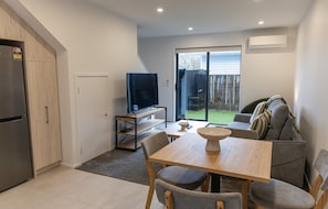 A compact kitchen with a built-in refrigerator, a dining area with a table and chairs, a television on a stand, and a sliding glass door leading to a backyard. - Stylish Two-Bedroom Townhouse Near Lower Hutt (Lower Hutt)