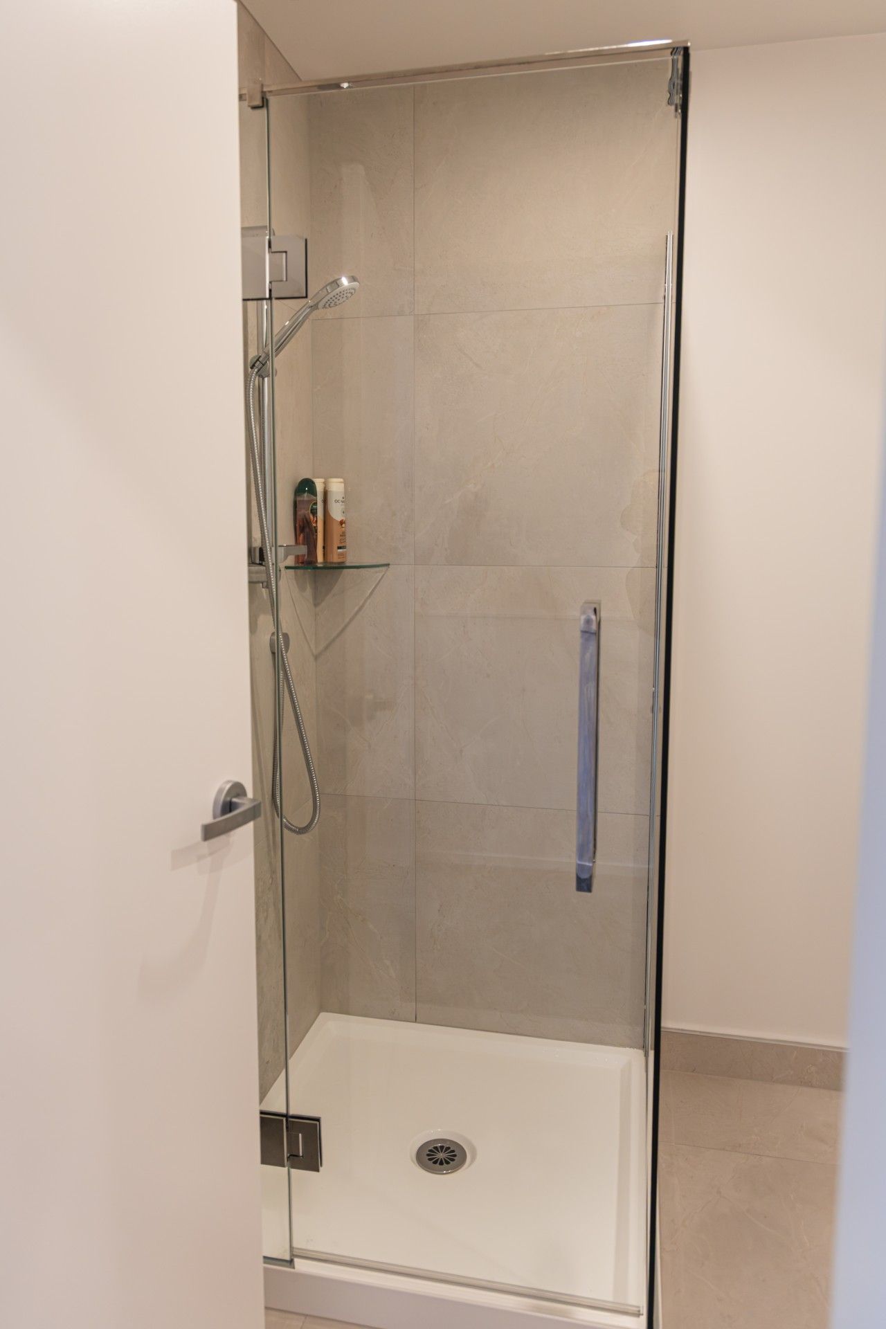 A glass-enclosed shower stall with a handheld showerhead, a handheld shower attachment, and a small shelf with toiletries.