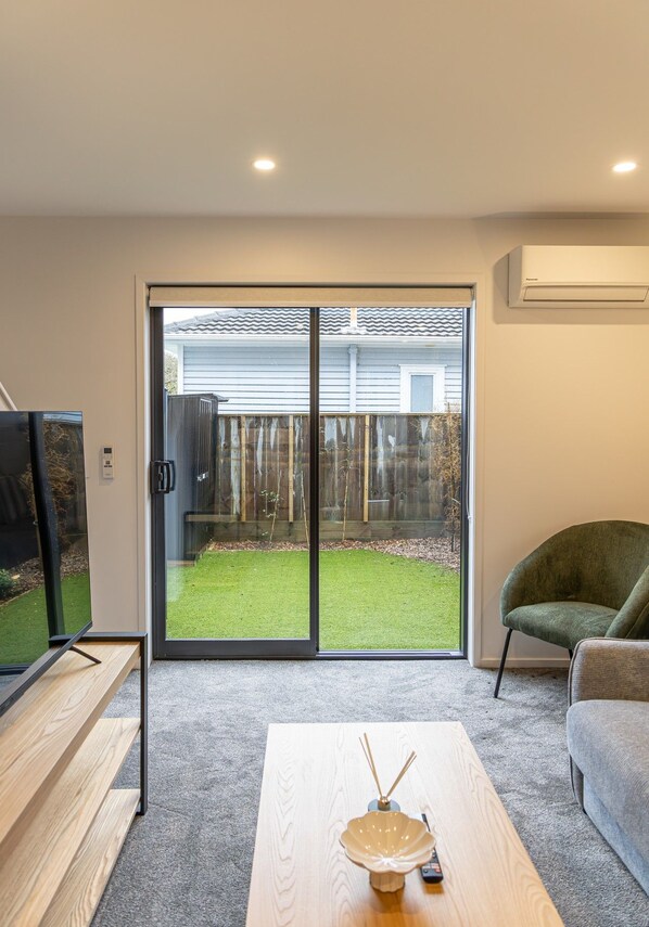 A modern living room with a sliding glass door leading to a backyard, a flat-screen TV, a wooden coffee table, and a green armchair.