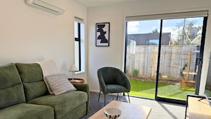 A living room with a green sofa, a wooden coffee table, a chair, and a sliding glass door leading to a backyard. - Modern 1BR Townhouse with Private Courtyard (Lower Hutt)
