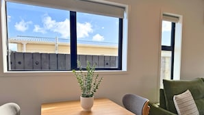 A room with a wooden table, two chairs, a green sofa, and a window with a view of a building and sky. - Modern 1BR Townhouse with Private Courtyard (Lower Hutt)