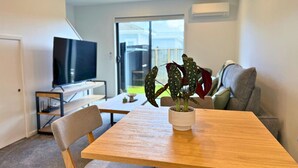 A modern living room with a wooden dining table, a grey sofa, a television, and a potted plant. - Bright & Spacious Wellington Townhouse (Lower Hutt)