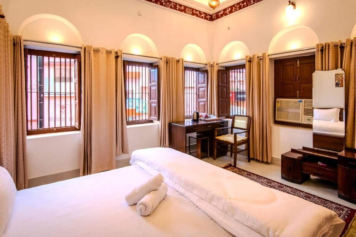 A hotel room with a large bed, a desk, and a chair. There are windows with curtains, a radiator, and a mirror.