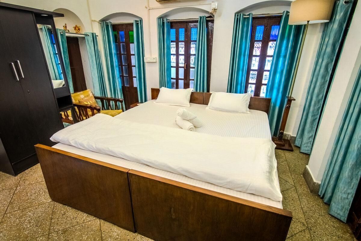 A large bed with a wooden frame and white bedding, flanked by two windows with blue curtains, a wooden chair, and a dark-colored wardrobe.
