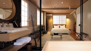 Une chambre d’hôtel moderne avec un plancher en bois, un grand lit, une salle de bain équipée d’un lavabo et d’un miroir, et une porte coulissante donnant sur une autre pièce.