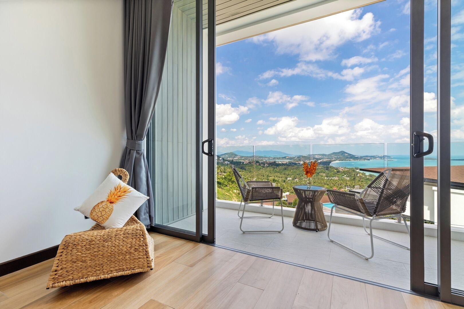 A balcony with a view of a coastal landscape, a wicker chair, and a small table with a vase.