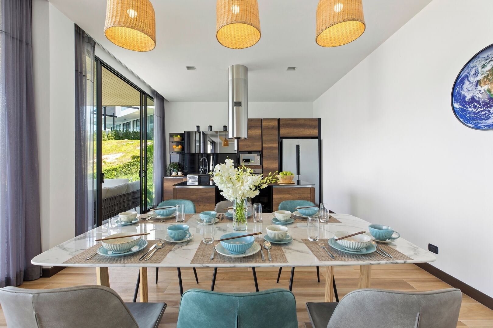 A modern dining area with a large table set for six, a kitchen in the background, and a large window with a view of greenery.