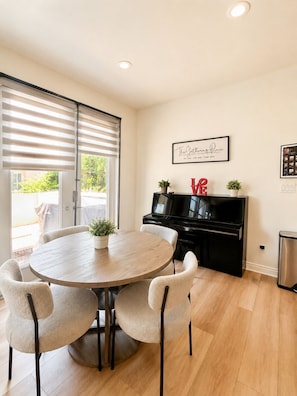 A dining area with a round table, four chairs, a piano, and a framed sign on the wall.