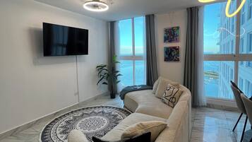 Una sala de estar moderna con un sofá, un televisor de pantalla plana, una alfombra estampada y vistas a un edificio y al agua a través de amplios ventanales.