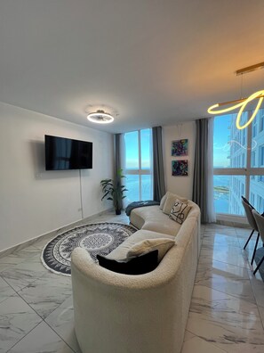 Ein modernes Wohnzimmer mit einer Couch, einem Flachbildfernseher, einem gemusterten Teppich und Blick auf ein Gebäude und Wasser durch große Fenster.