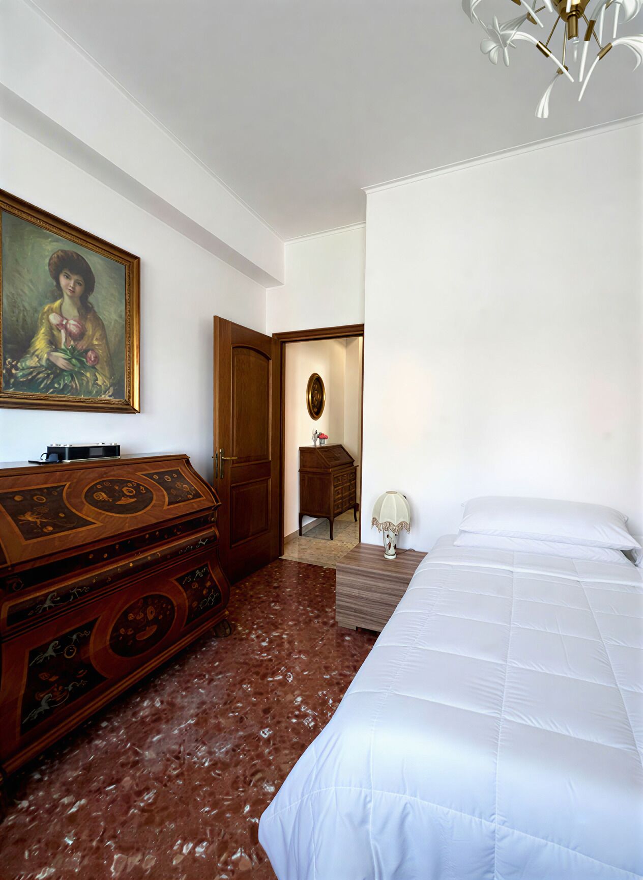 A room with a bed, a dresser, a painting, a door, and a chandelier.