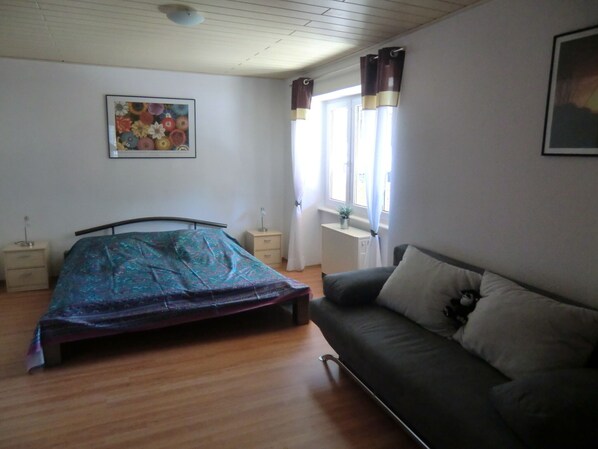 A bedroom with a bed, a sofa, a nightstand, a window with curtains, and a wall-mounted picture.