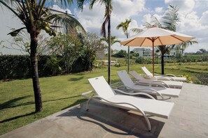 Zona de piscina amb gandules i una ombra, envoltada de palmeres i un jardí ben cuidat.