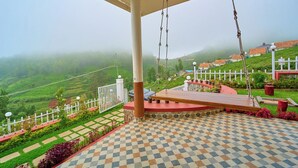 A terrace with a checkered floor, a swing, a white fence, and a view of green hills. - Emy Misty Charm Villa Ooty by VOYE HOMES (Ootacamund)