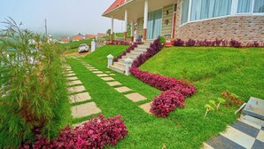 A house with a red roof, a front porch, and a well-maintained lawn with a stone pathway. - Emy Misty Charm Villa Ooty by VOYE HOMES (Ootacamund)