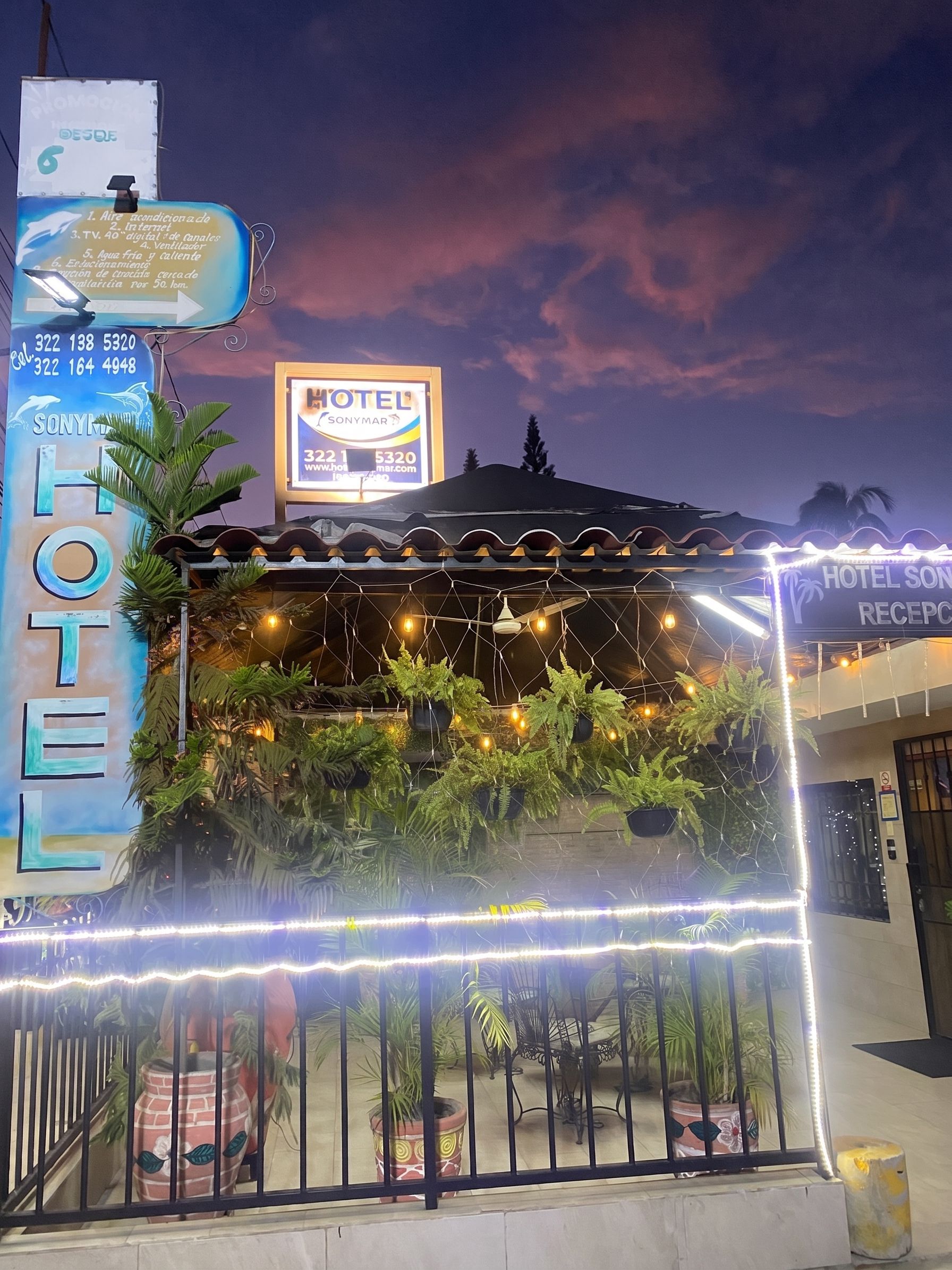 A hotel with a signboard, a reception area with plants, and a fence.