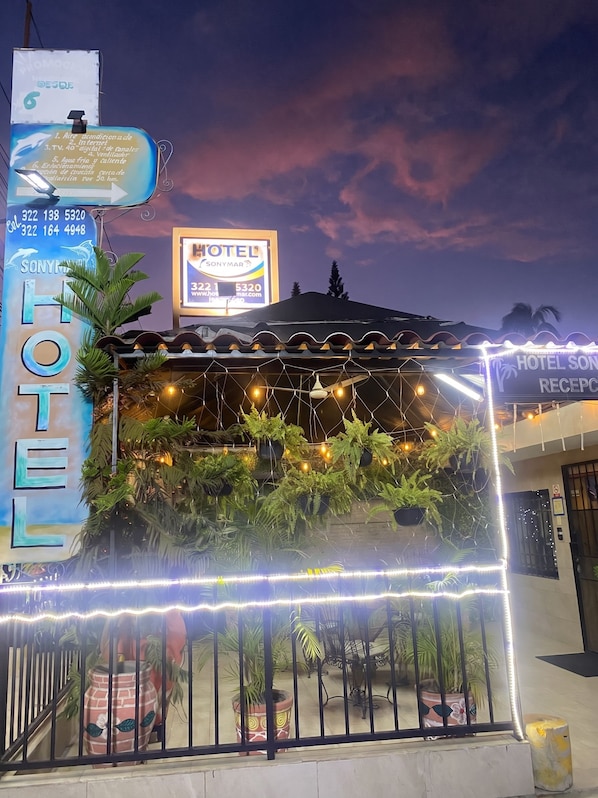 A hotel with a signboard, a reception area with plants, and a fence.
