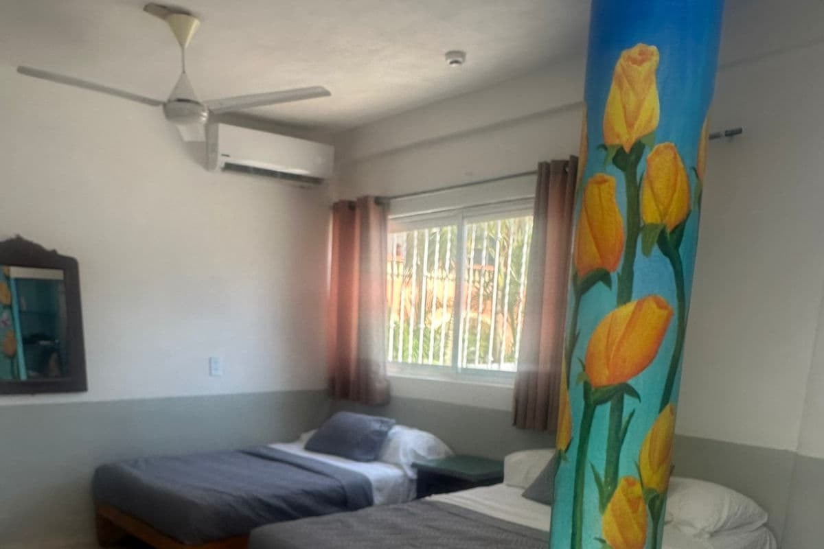 A hotel room with two beds, a ceiling fan, a window with curtains, and a pillar with a floral design.