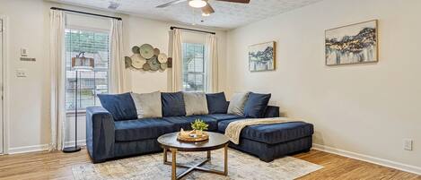 A living room with a dark blue sectional sofa, a round coffee table, a ceiling fan, and artwork on the walls.