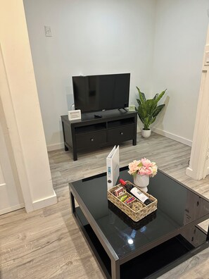 A modern living room with a flat-screen TV, a dark wooden entertainment center, a glass coffee table with a basket, and a potted plant.