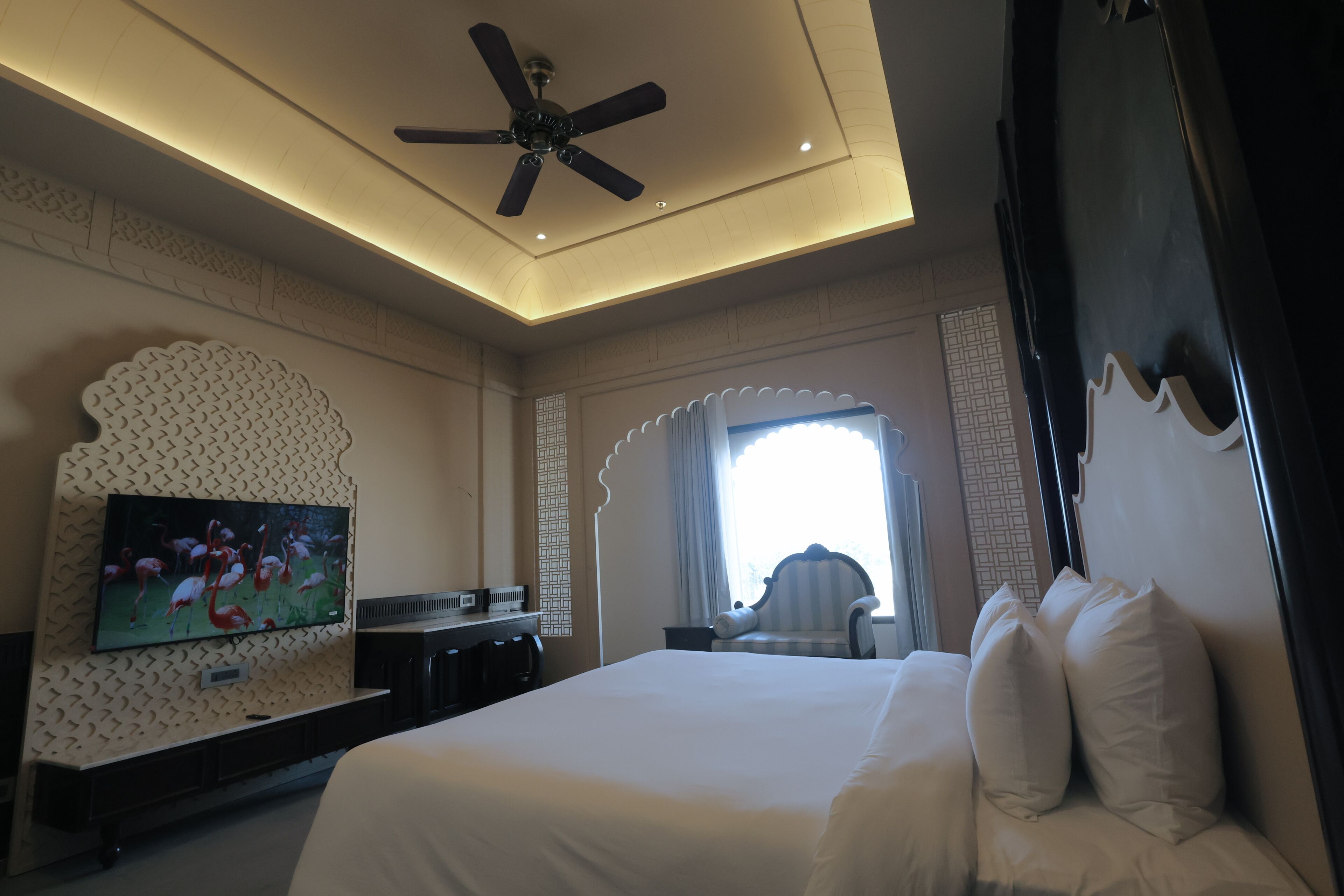 A hotel room with a bed, television, ceiling fan, and a window with curtains.