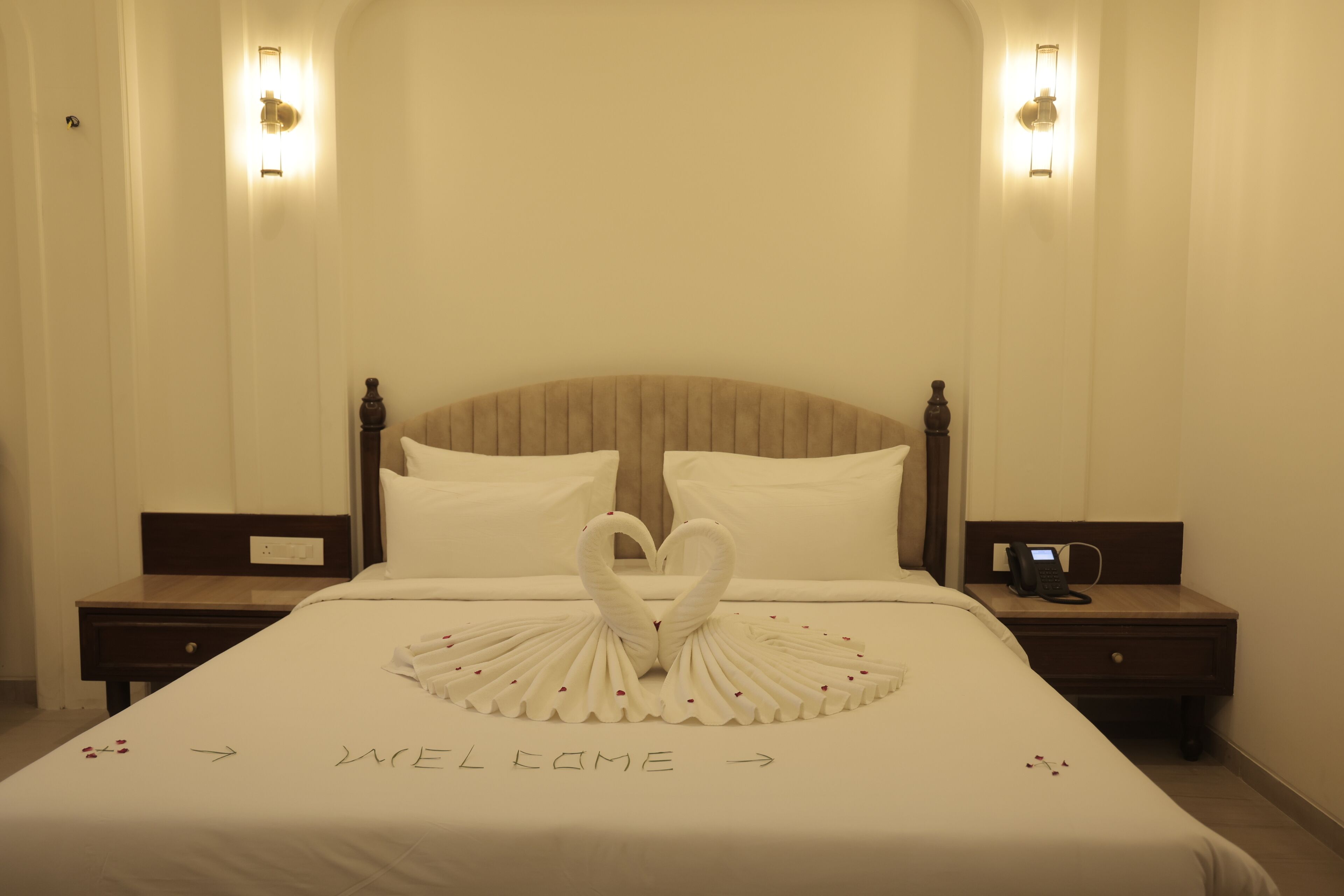 A hotel room with a bed featuring a heart-shaped towel arrangement and 'WELCOME' written on the bedspread.