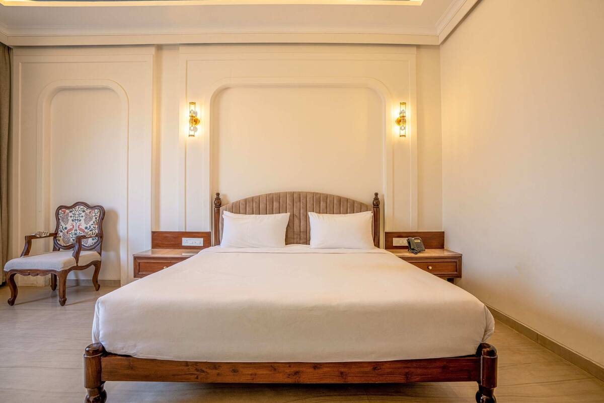 Habitación de hotel ordenada con una cama grande, dos mesitas de noche, una silla e iluminación en la pared.