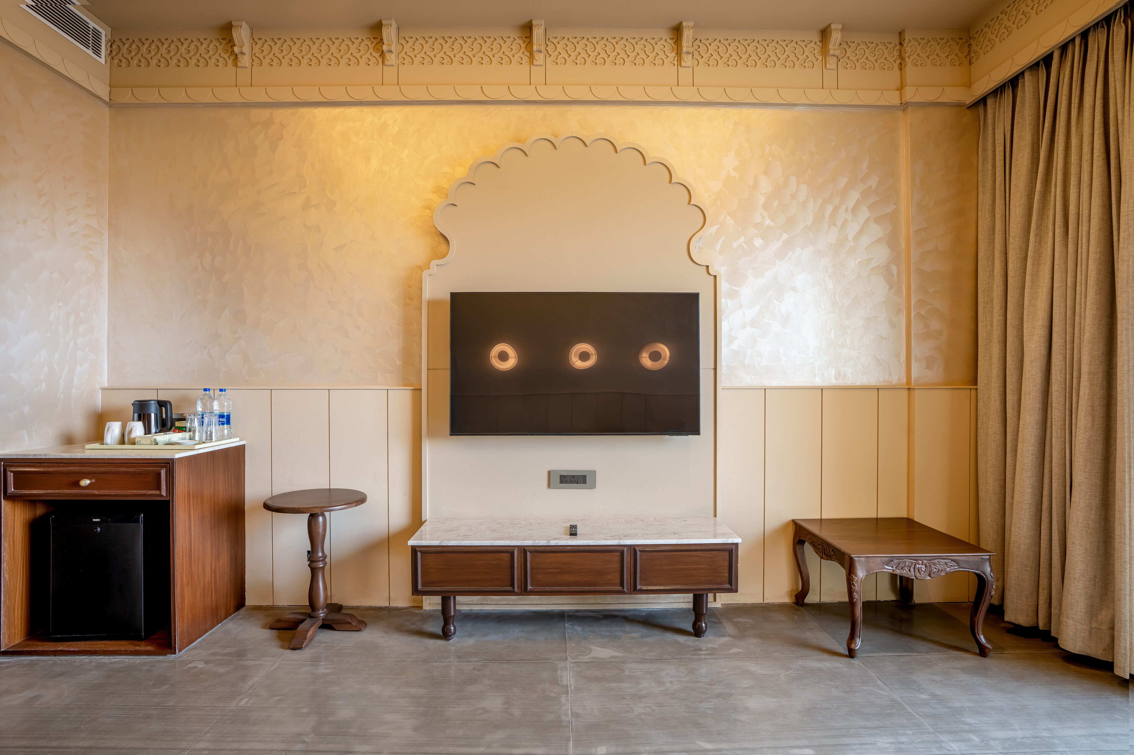 A hotel room with a TV mounted on the wall, a small cabinet, a round table, and a decorative alcove.